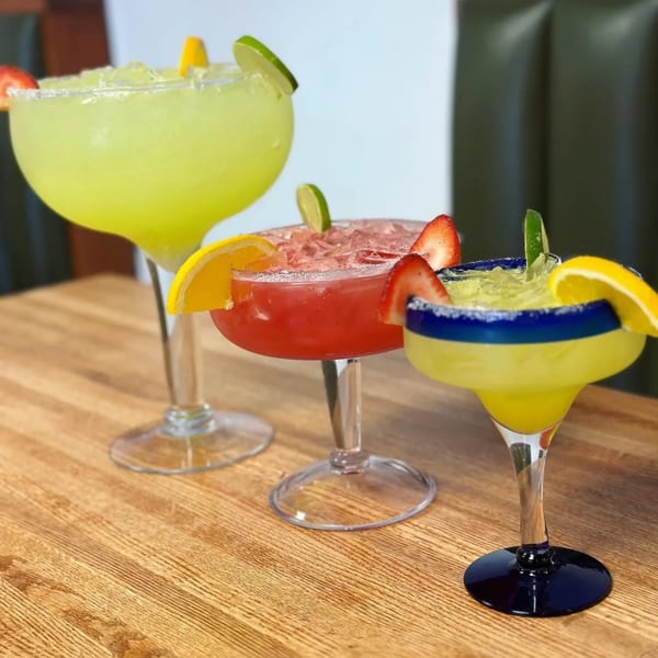Three colorful margaritas, garnished with fruit and salt, on a wooden table at Burger Taco.